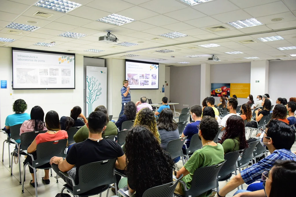 Vista de trás de uma plateia em uma sala de conferência, assistindo a um palestrante que usa duas telas de projeção.
