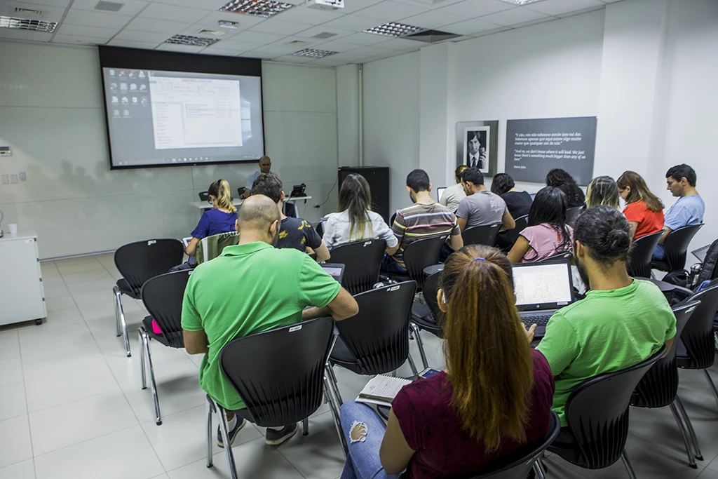 Vista de trás de uma sala de aula cheia de pessoas assistindo a uma apresentação em uma tela de projeção.