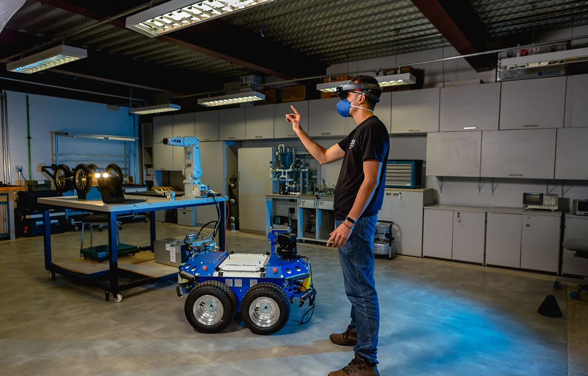 Homem usando óculos de realidade virtual e máscara em laboratório, apontando para um robô de quatro rodas que está no chão.