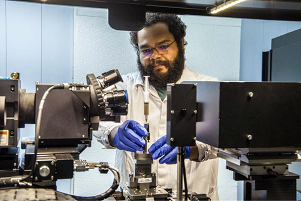 Homem de jaleco branco, usando luvas azuis, operando um equipamento de análise complexo em um laboratório.