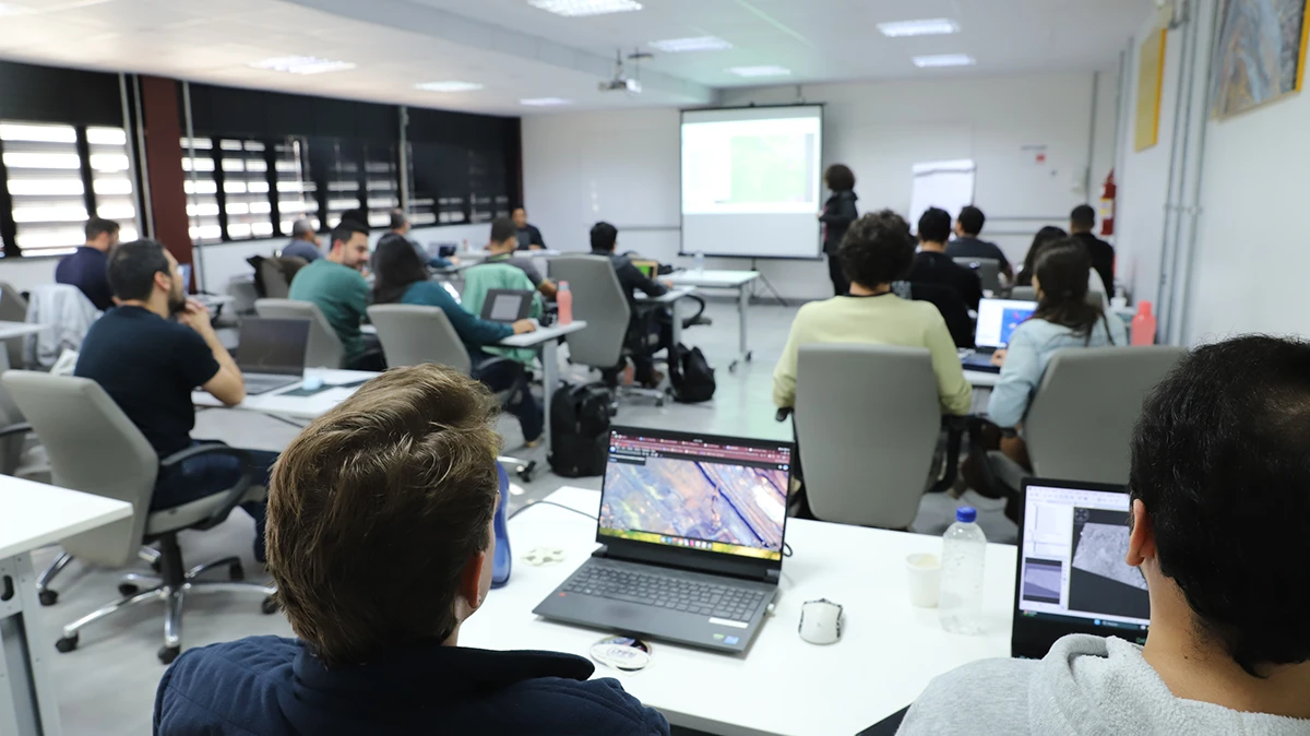Vista de trás de um grupo de alunos em uma sala de aula com notebooks, assistindo a uma apresentação projetada em uma tela.